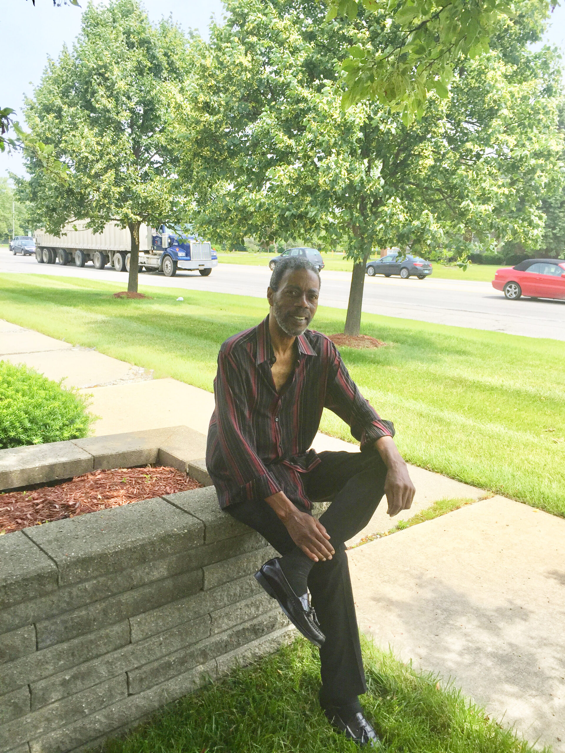 Man sitting on a low stone wall in a sunny suburban area.
