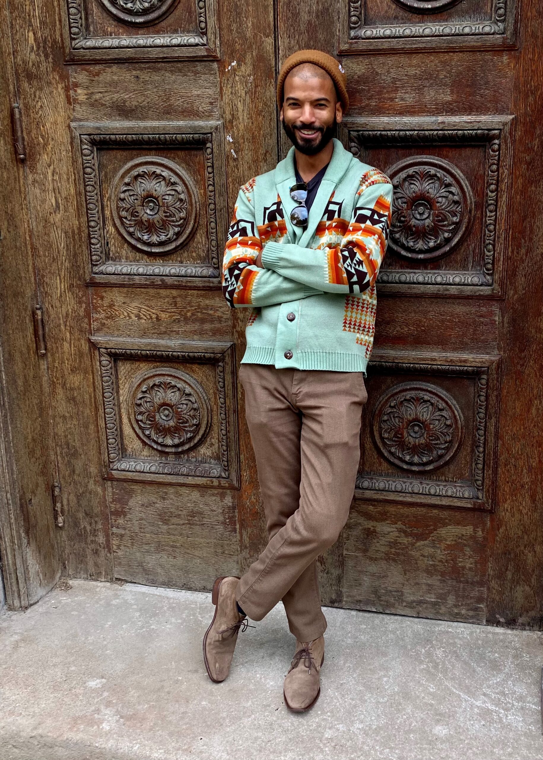 A man in a patterned cardigan and brown beanie stands against a carved wooden door.