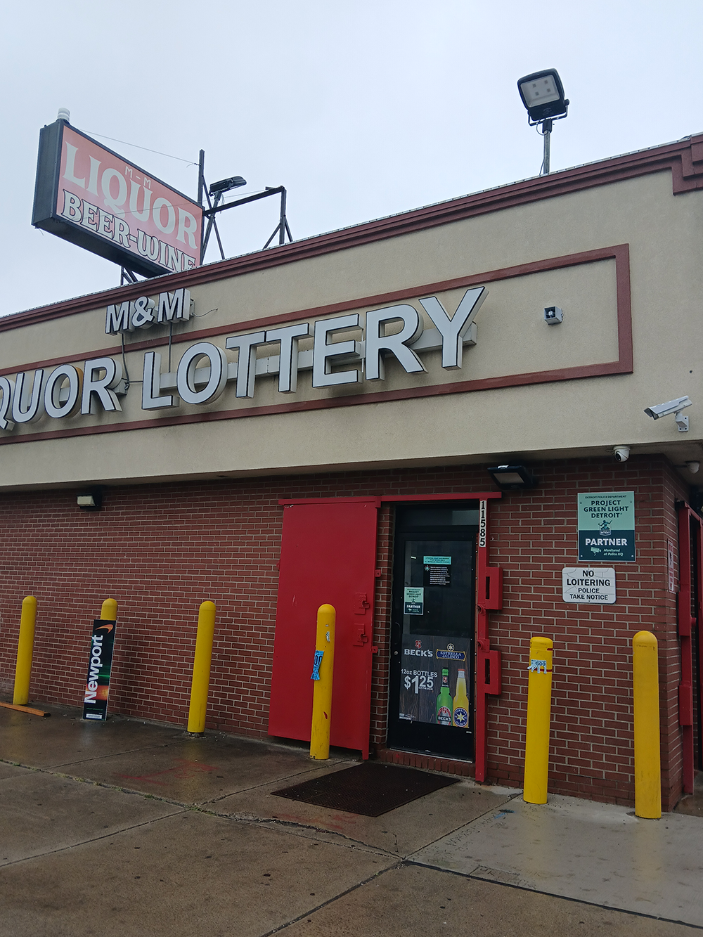 Exterior of a liquor and lottery store with a red door and yellow bollards.