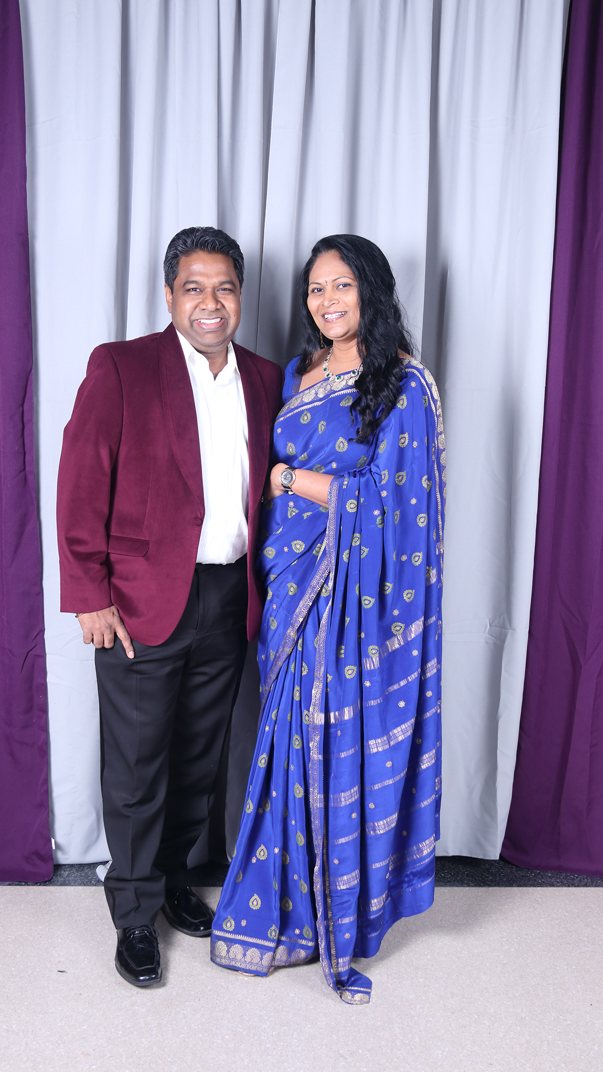 A couple poses together in formal attire, with the woman in a blue saree and the man in a maroon blazer.